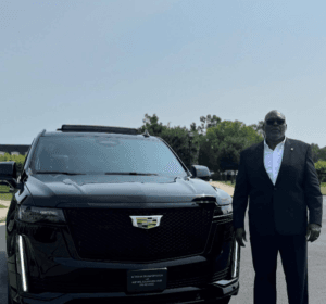 Person in a dark suit with sunglasses standing beside a shiny black Cadillac SUV on a paved area, with greenery and clear sky in the background.