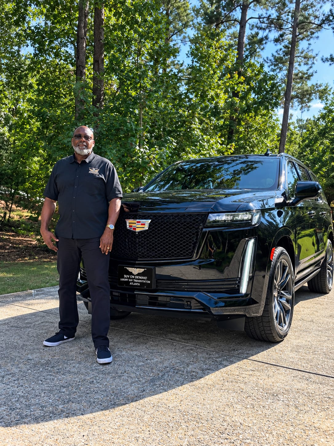 04278365-FD05-4DAF-A309-5970C25FC2FB Person in dark clothing posing beside a shiny black SUV on a sunny driveway, with tall green trees in the background.